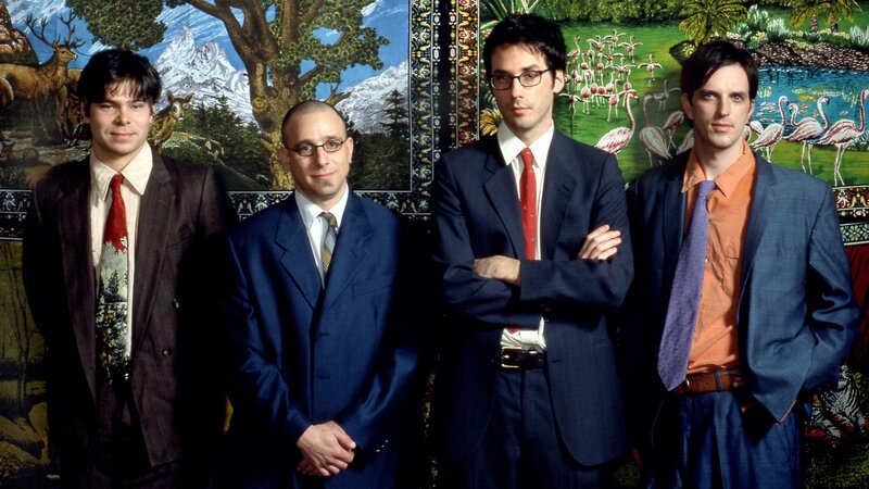 Four men in suits pose in front of a tapestry