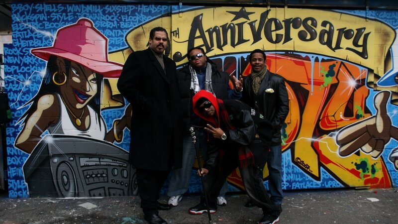 Four men pose in front of graffiti