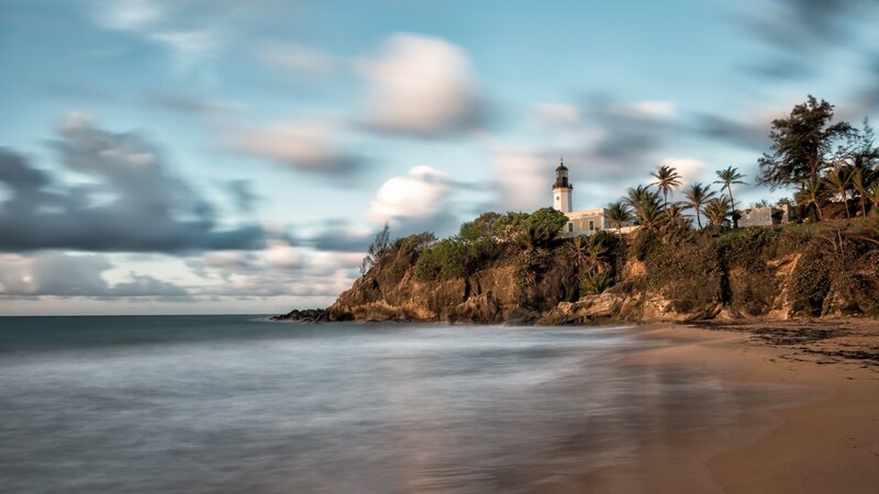 Lighthouse on Rocky Cliff — Blue, Serene, Sunset, Photograph, Ocean, Beach, Palm Trees