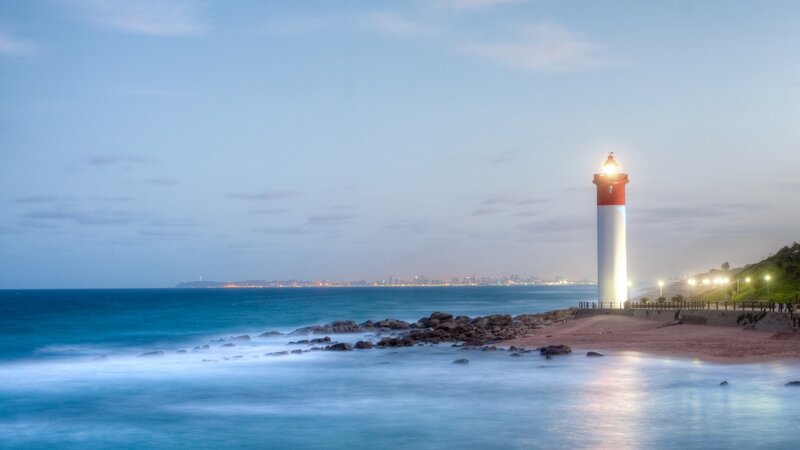 Lighthouse on Rocky Shore — Blue, Serene, Sunset, Photograph, Ocean, City Skyline