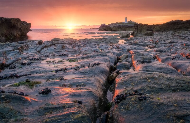 Lighthouse on Rocky Shore — Orange, Serene, Sunset, Photograph, Sunrise