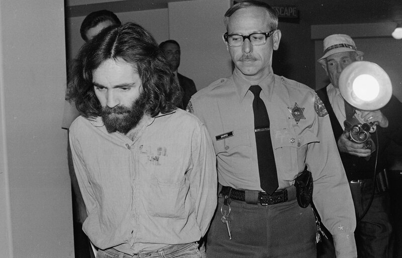 Man with long hair escorted by officer