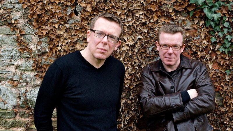 Two men standing by a leafy wall